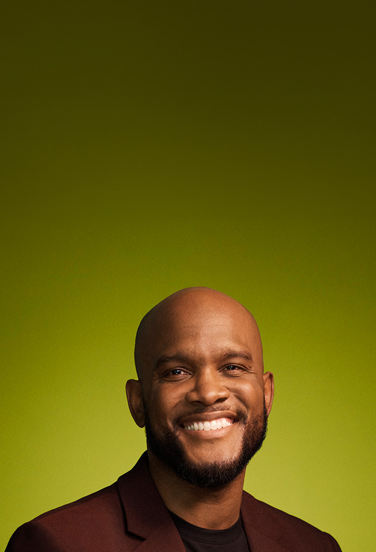 Headshot of bald Black man in his 30s, smiling, wearing a maroon blazer, on a green background