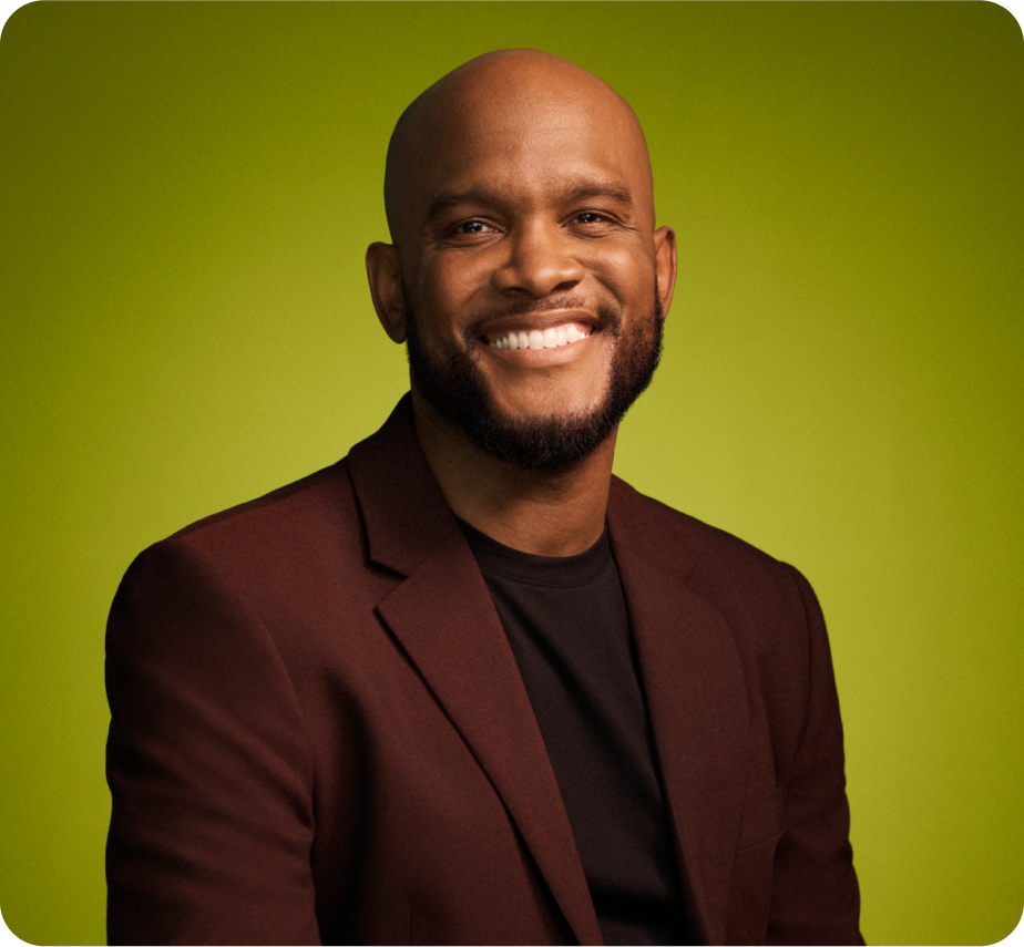 Headshot of bald Black man, smiling, wearing a maroon blazer, on a green background